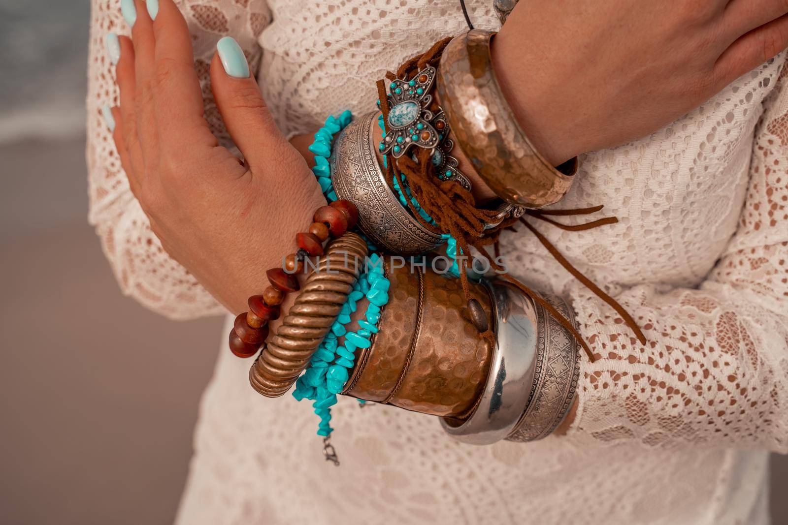 Model in boho style in a white long dress and silver jewelry on the beach. Her hair is braided, and there are many bracelets on her arms