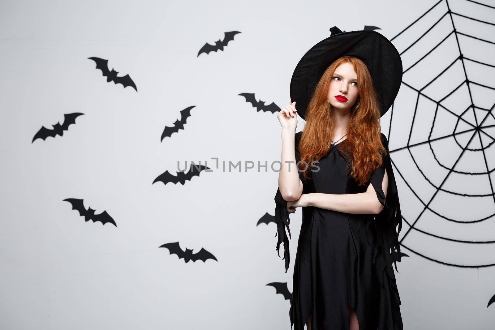 Halloween witch concept - Halloween Witch holding posing with serious expression over dark grey studio background with bat and spider web. by Benzoix