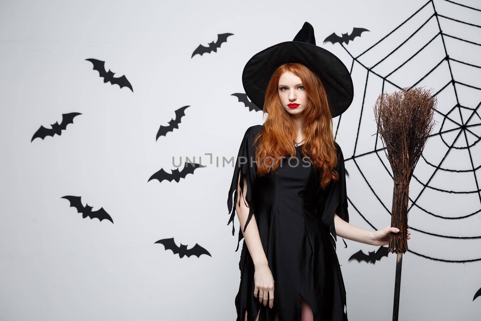 Halloween Witch Concept - Portrait of beautiful young witch with broomstick over grey wall with bat and spider web background