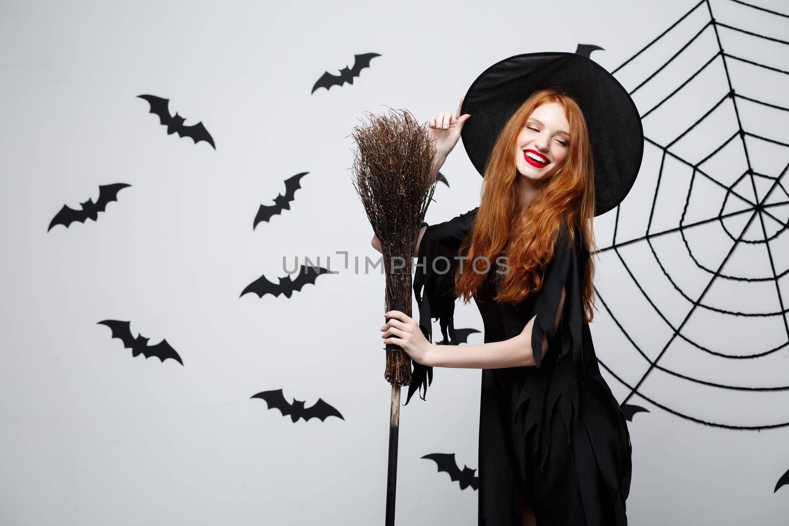 Halloween Witch Concept - Portrait of beautiful young witch with broomstick over grey wall with bat and spider web background. by Benzoix