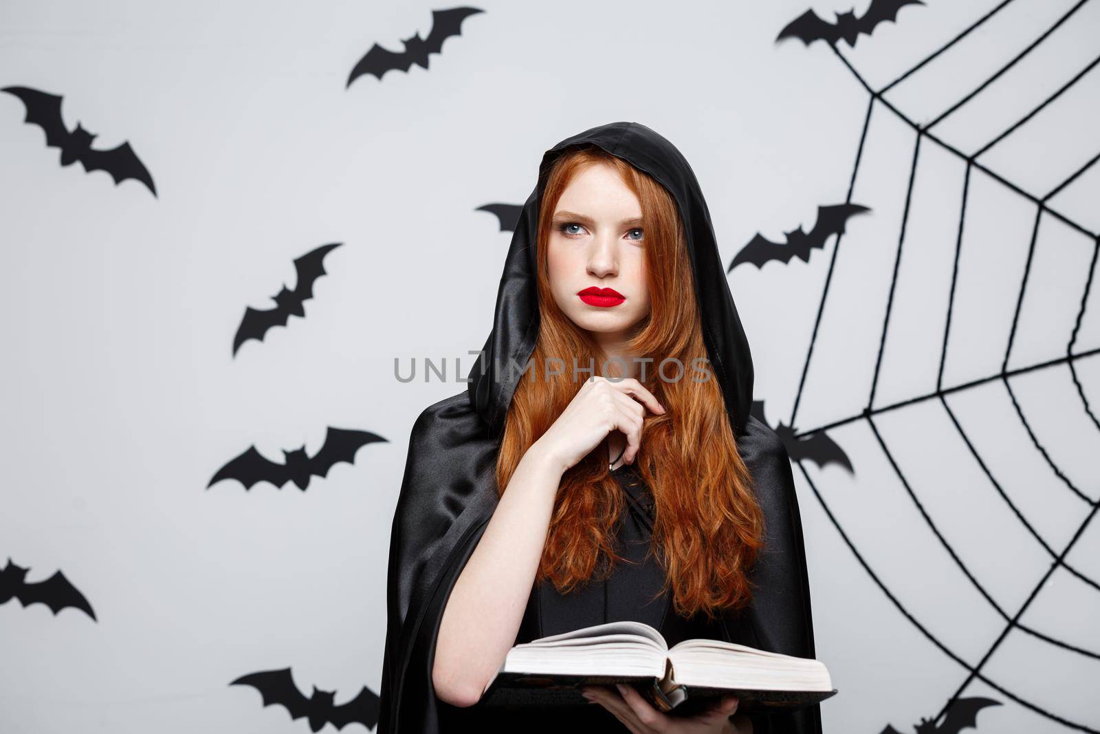 Halloween Concept - Beautiful dark priest casting spell with magic book on grey background. by Benzoix