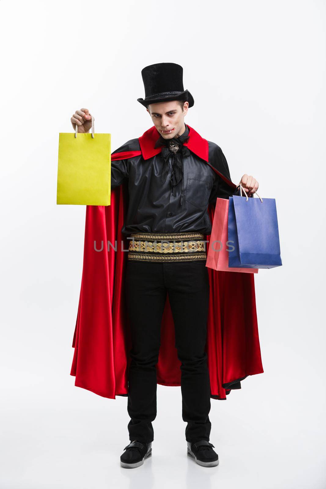 Vampire Halloween Concept - Happy handsome caucasian Vampire holding colorful shopping bag on white background. by Benzoix