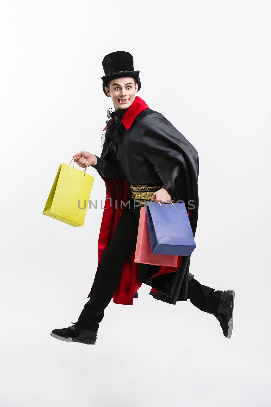 Vampire Halloween Concept - Happy handsome caucasian Vampire holding colorful shopping bag on white background. by Benzoix