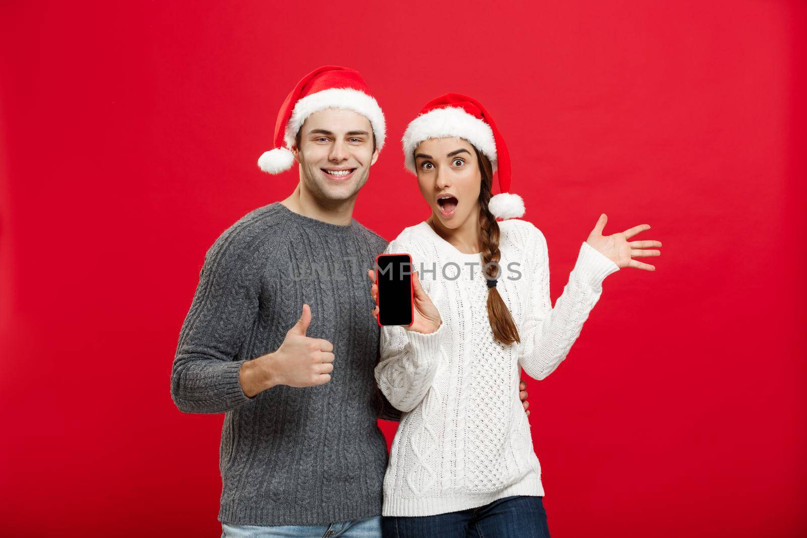 Christmas concept - Happy young couple in christmas sweaters showing thump up gesture with mobile phone.