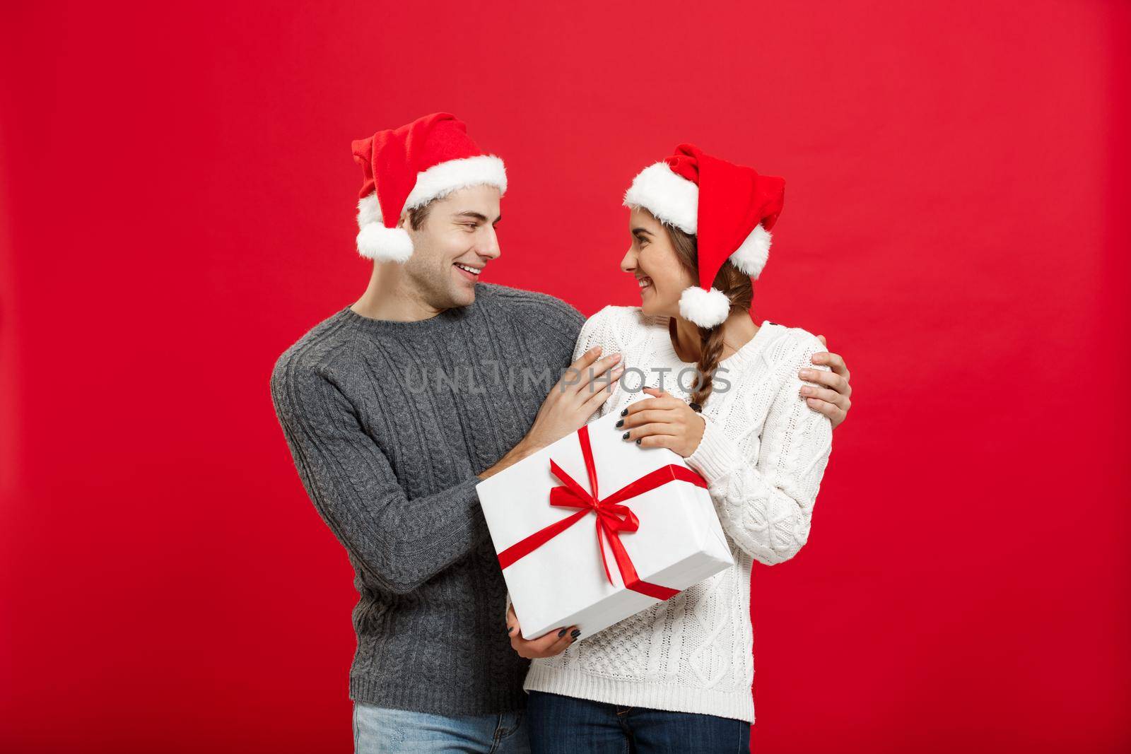 Christmas Concept - Young woman covering man's eyes with hand and giving surprise big gift. Isolated on Red background.