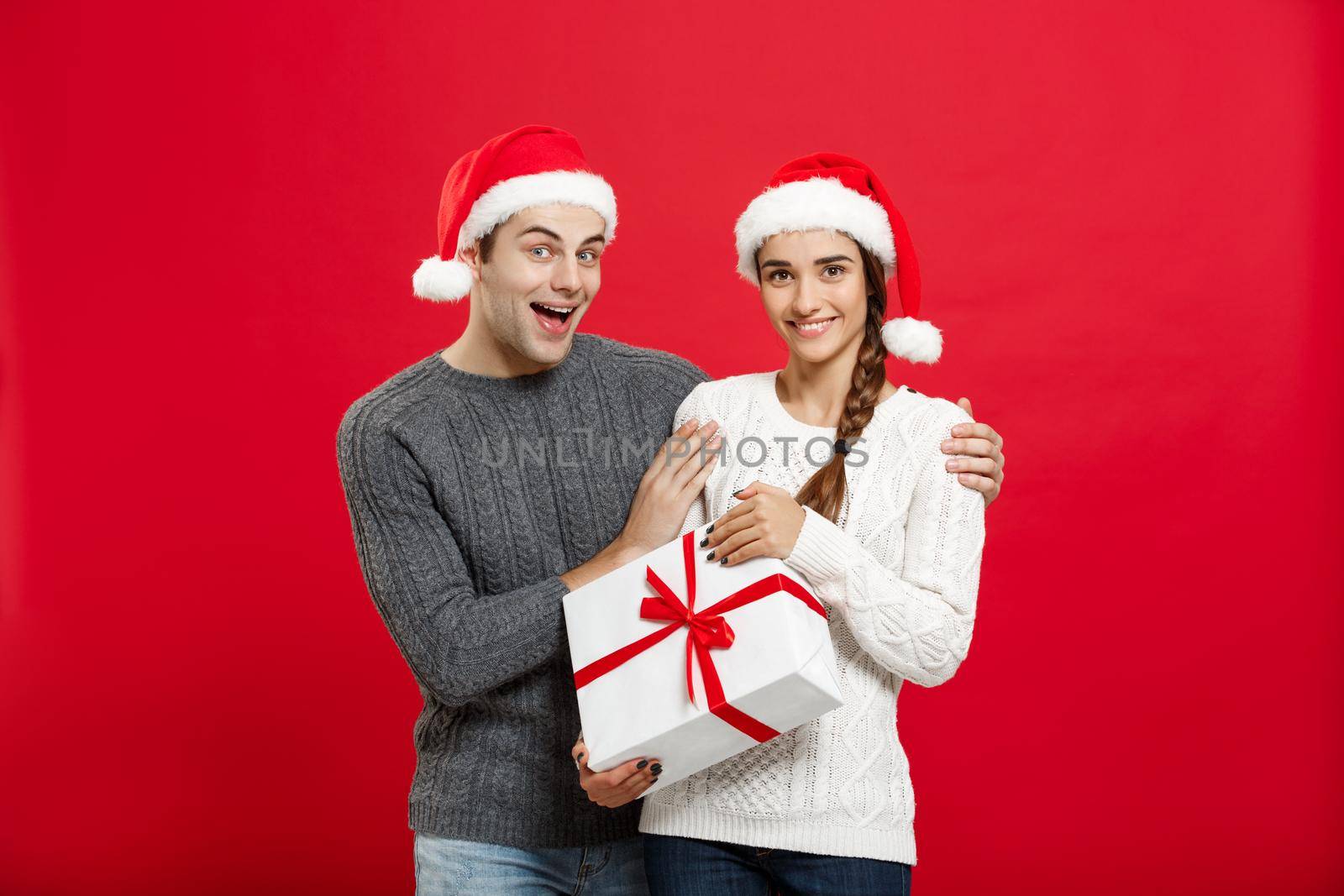 Christmas Concept - Young woman covering man's eyes with hand and giving surprise big gift. Isolated on Red background by Benzoix