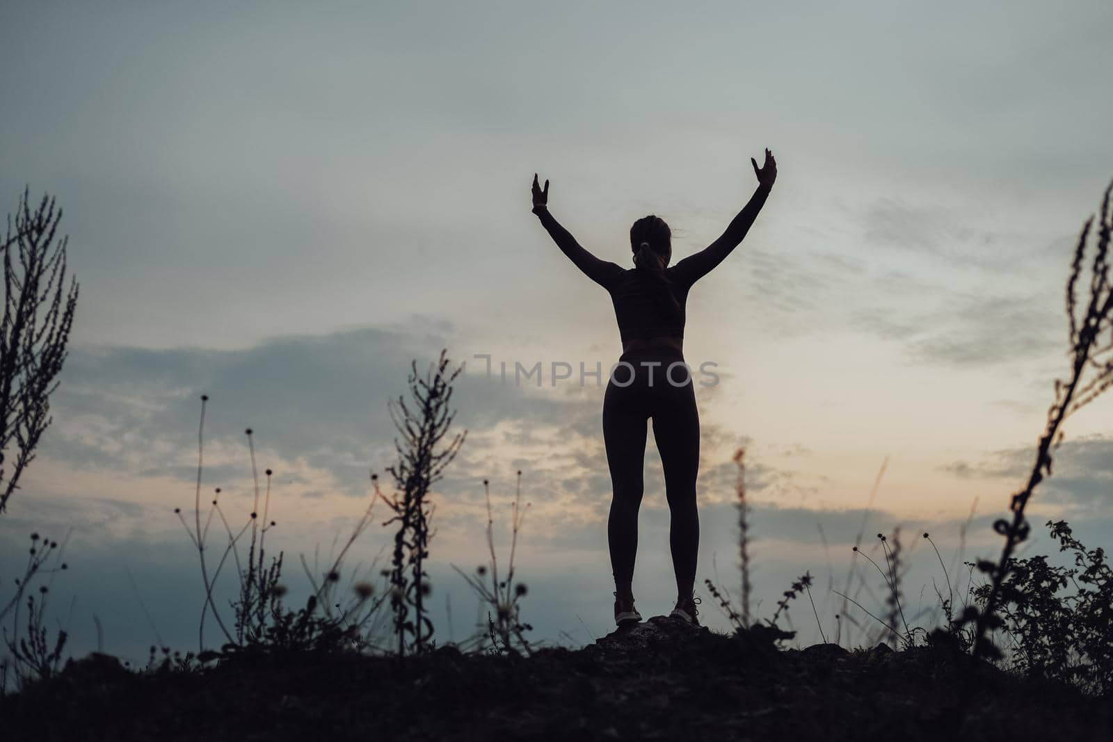 Silhouette of Sporty Young Woman Standing on Top of Hill and Raised Hands Up at Evening