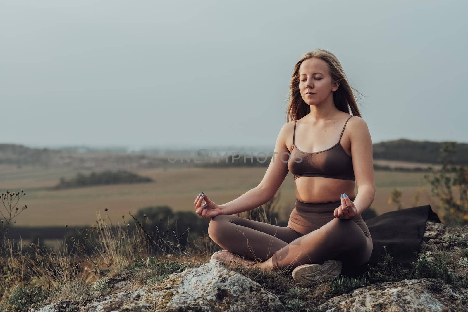 Sporty Girl Sitting in Lotus Pose and Doing Meditation Exercises on Top of a Hill, Young Woman Practice Yoga Outdoors with Panoramic Landscape at Sunset by Romvy