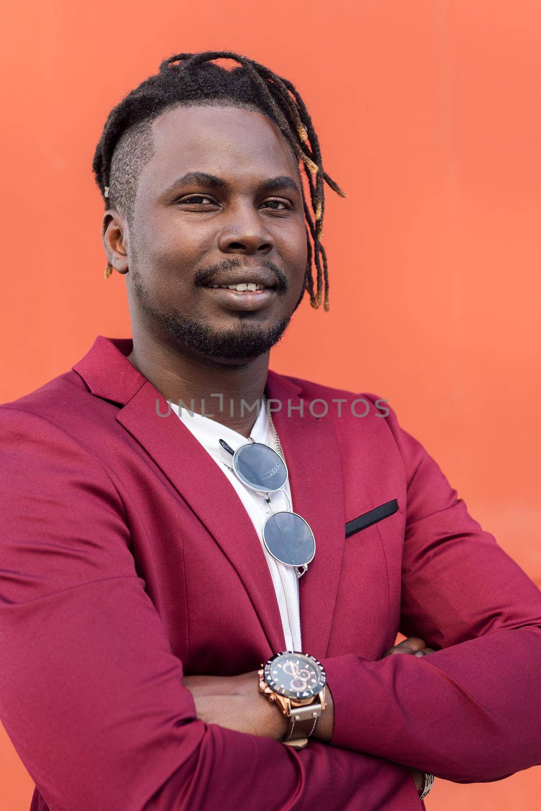 vertical portrait of a stylish black businessman posing looking at camera with arms crossed in front of red background, copy space for text