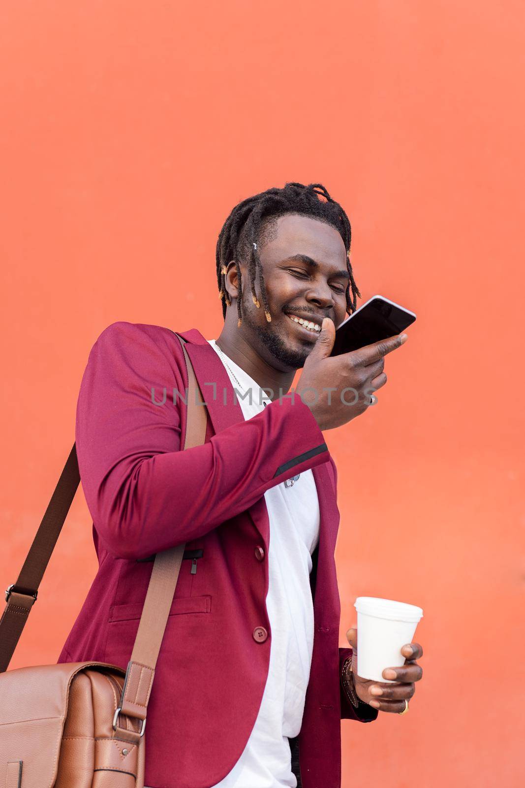 smiling african business man dressed in suit with briefcase and coffee records a voice message on his phone in front of a red background, copy space for text, concept of technology and communication