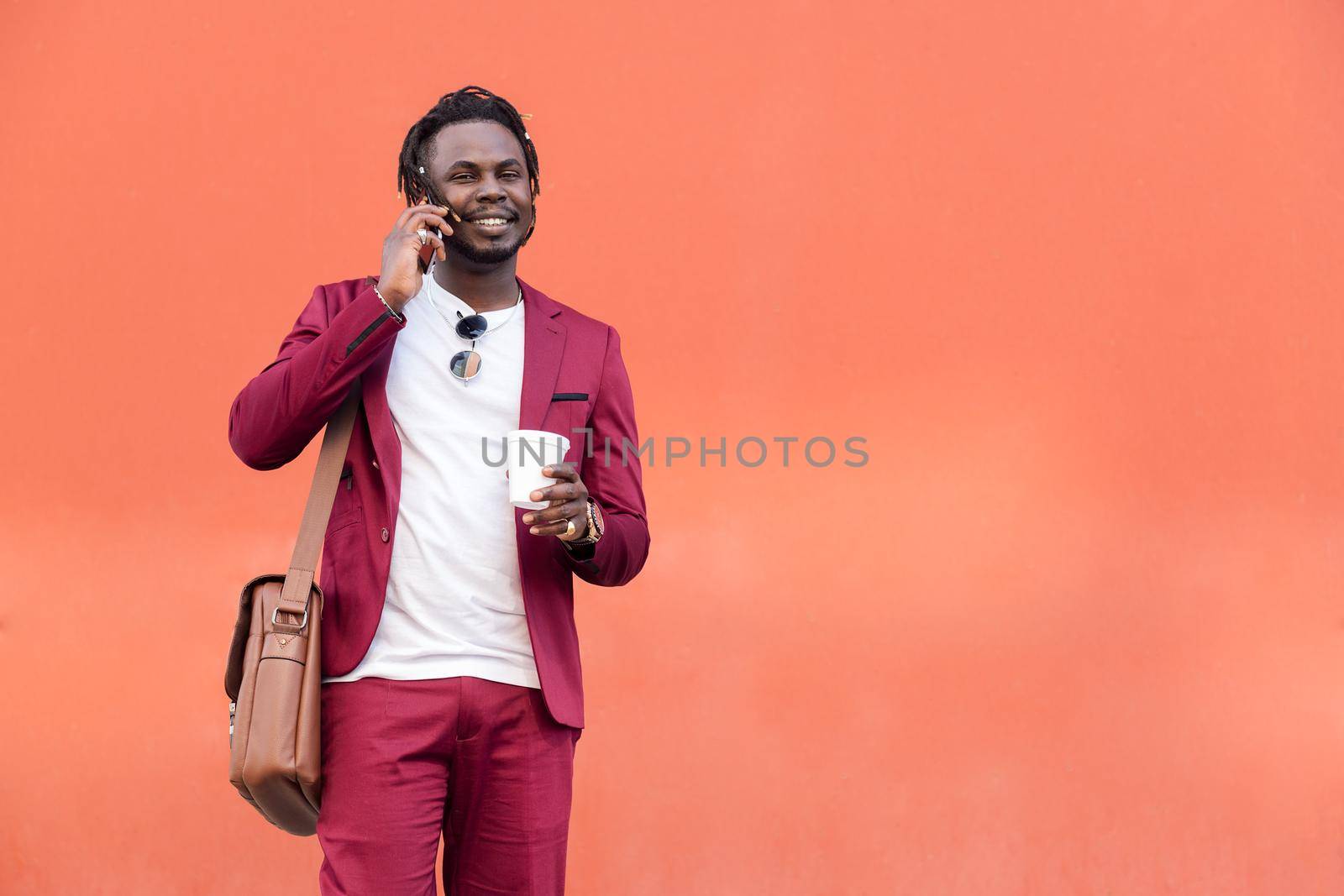 stylish black businessman dressed in suit with briefcase and coffee talks on smart phone in front of red background, copy space for text, concept of technology and communication