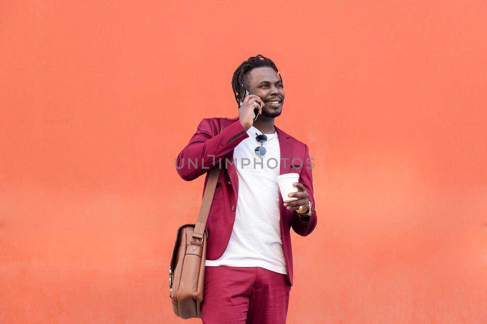 stylish african businessman dressed in suit smiles with a coffee on his hand while talks by phone in front of red background, copy space for text, concept of technology and communication