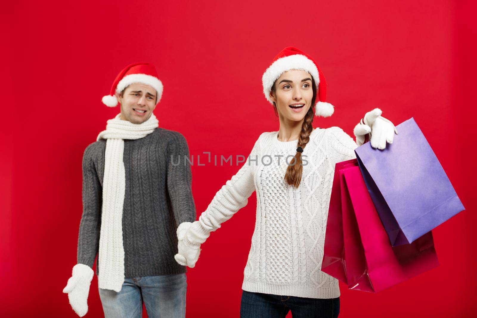 Christmas Concept - Beautiful girlfriend force her boyfriend to go shopping over red christmas background.