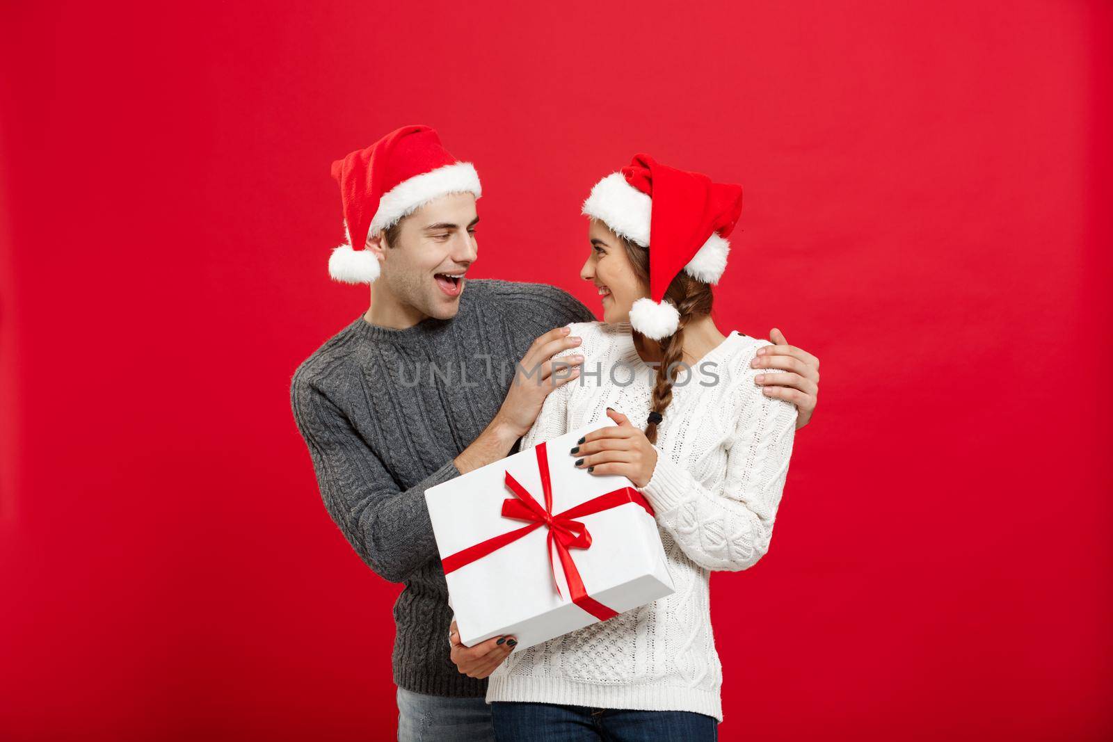Christmas Concept - Young woman covering man's eyes with hand and giving surprise big gift. Isolated on Red background by Benzoix