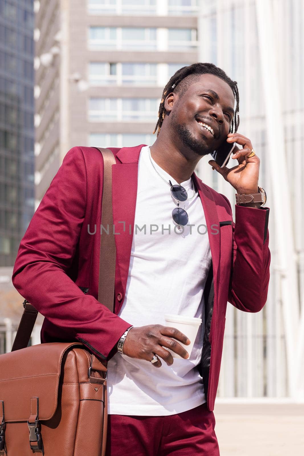 vertical portrait of a smiling african businessman walking through the city financial downtown talking on the phone with a coffee in his hand, concept of technology and communication