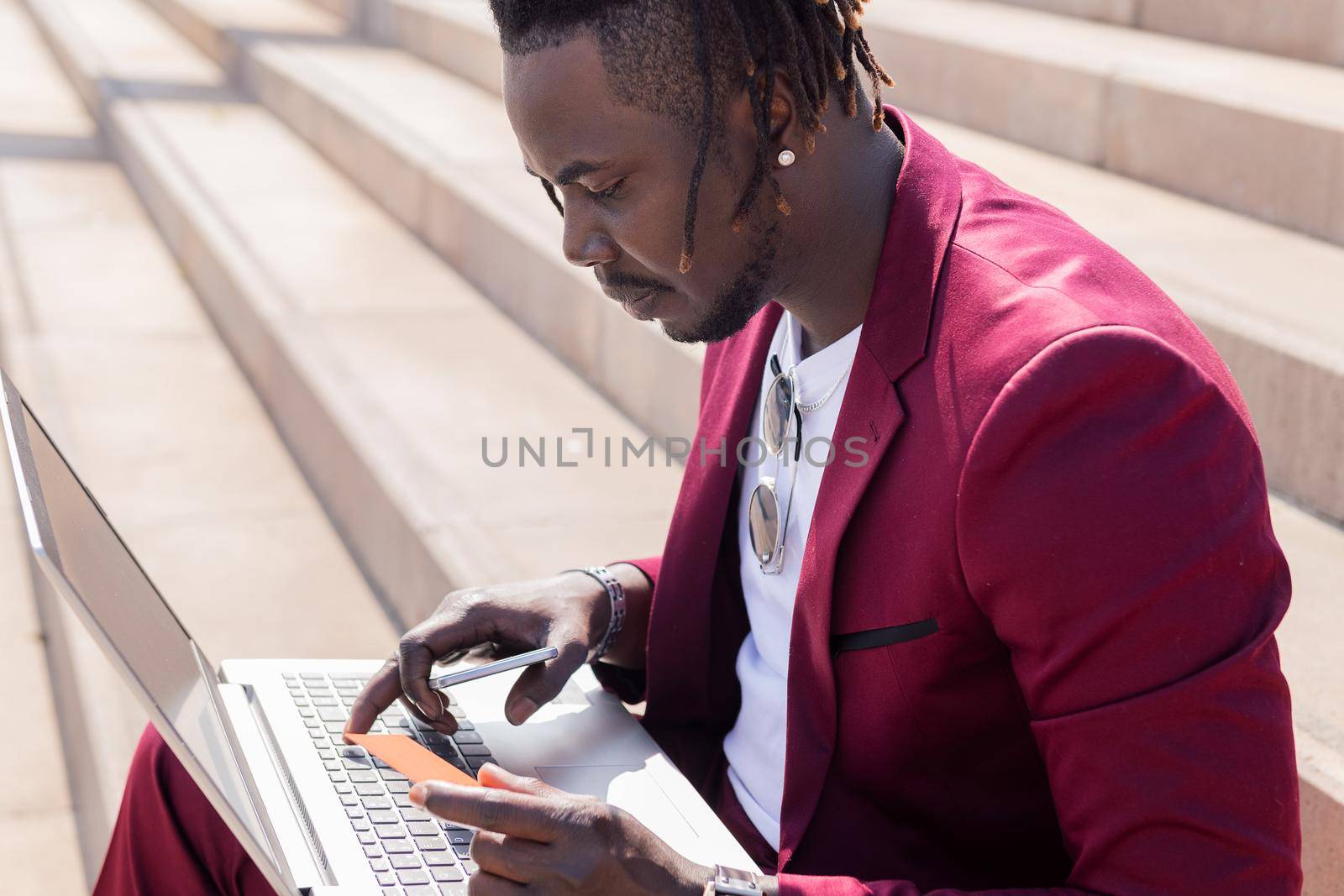elegant black man sitting outdoors pays with his credit card for the online purchase done on his laptop computer, technology and online shopping concept