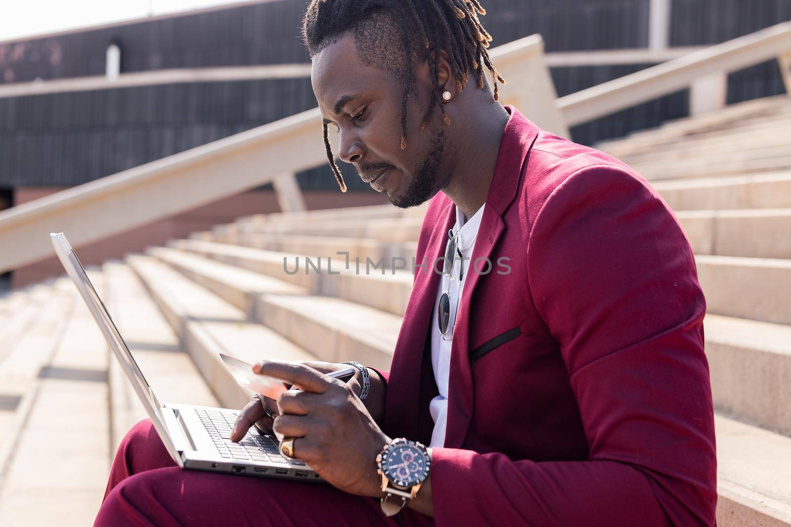 stylish african man sitting outdoors pays with his credit card for the online purchase done on his laptop computer, technology and online shopping concept