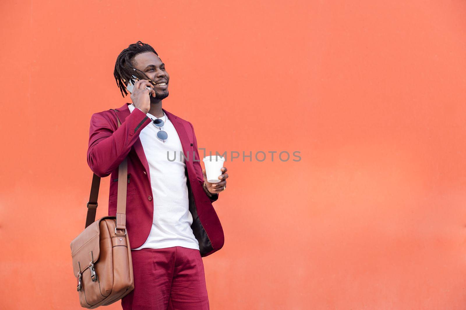 stylish african businessman dressed in suit with briefcase and coffee talks on smart phone in front of red background, copy space for text, concept of technology and communication