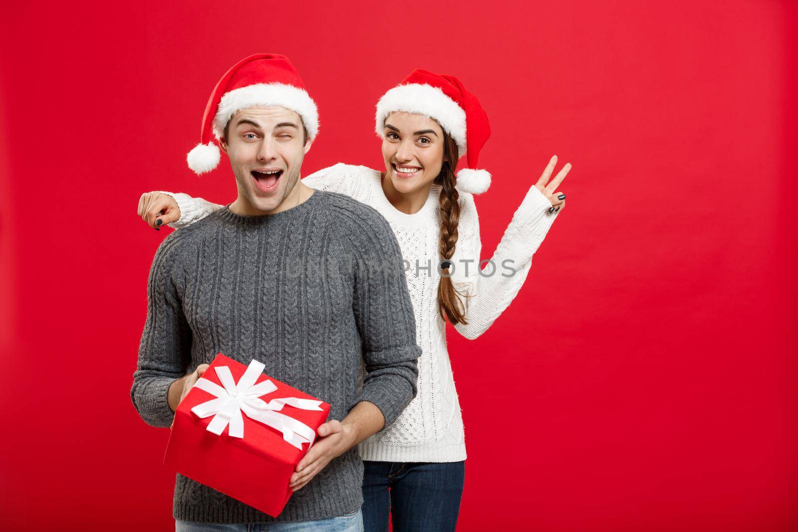 Christmas Concept - Young woman covering man's eyes with hand and giving surprise big gift. Isolated on Red background by Benzoix