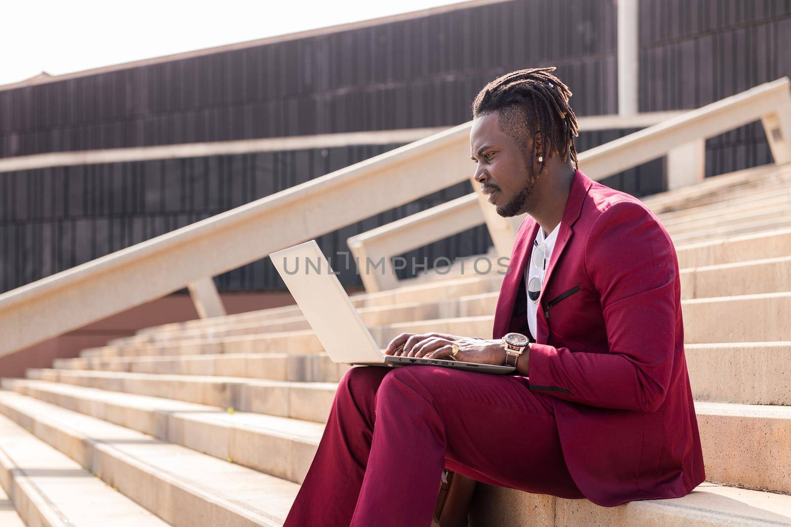 stylish black businessman works with his laptop sitting on a staircase in the city, technology and remote work concept