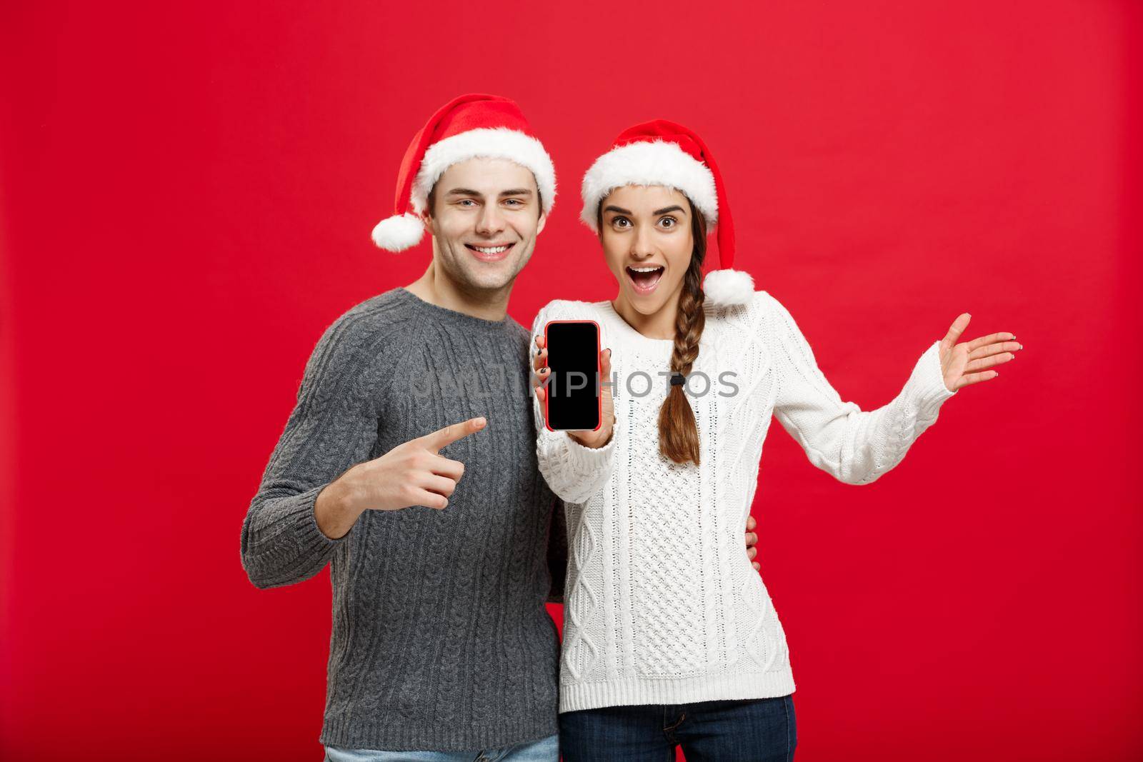 Christmas concept - Happy young couple in christmas sweaters pointing finger with mobile phone by Benzoix