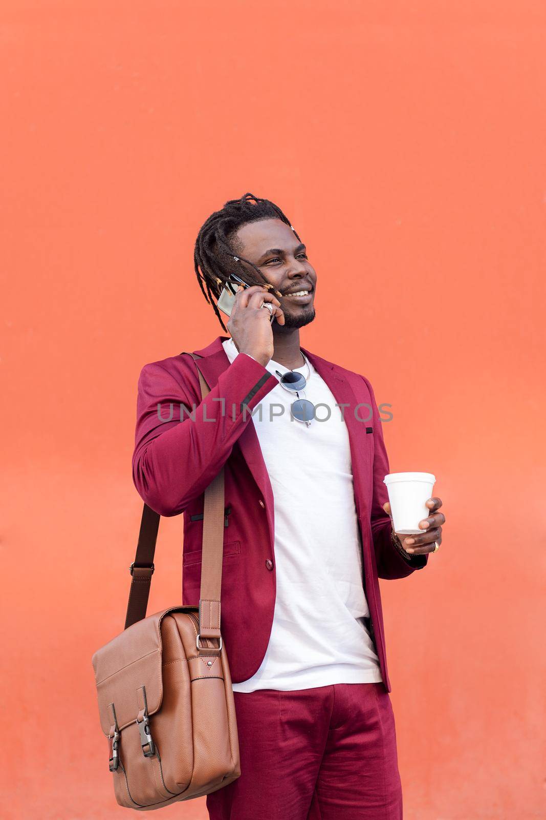 vertical photo of a stylish black businessman dressed in suit with briefcase and coffee talks on smart phone in front of red background, copy space for text, concept of technology and communication