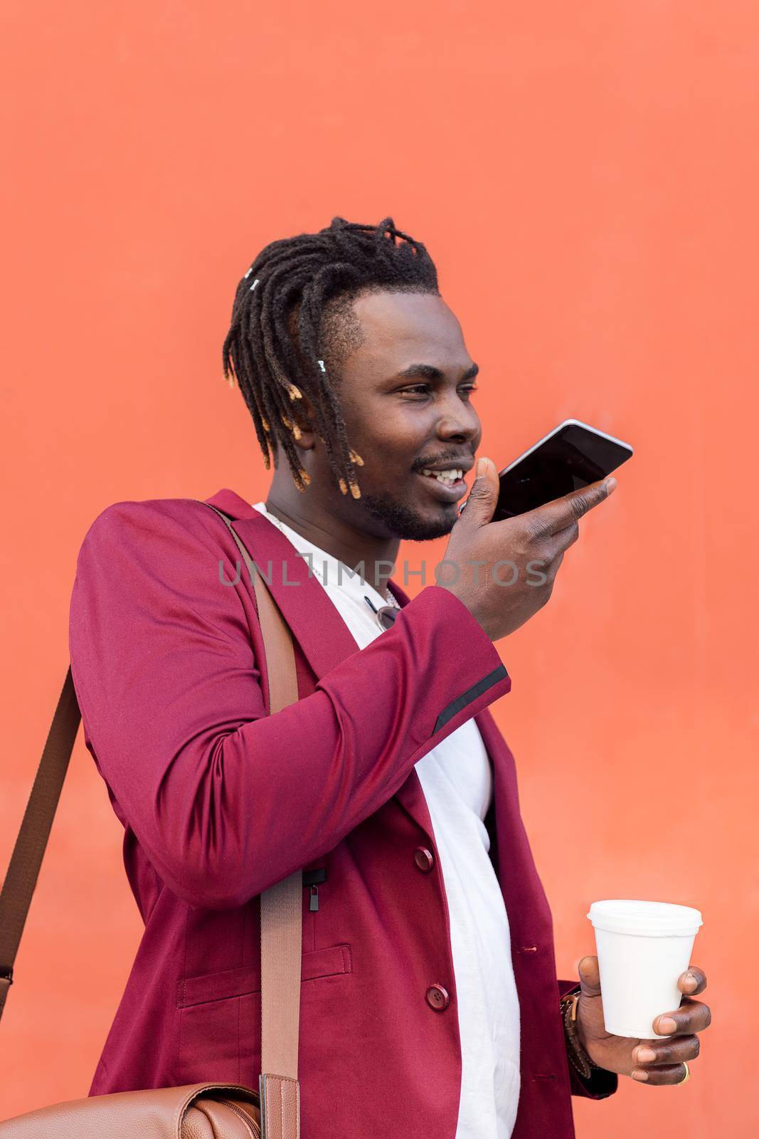 vertical portrait of a stylish black businessman dressed in suit and holding a coffee records voice message on phone in front of red background, concept of technology and communication