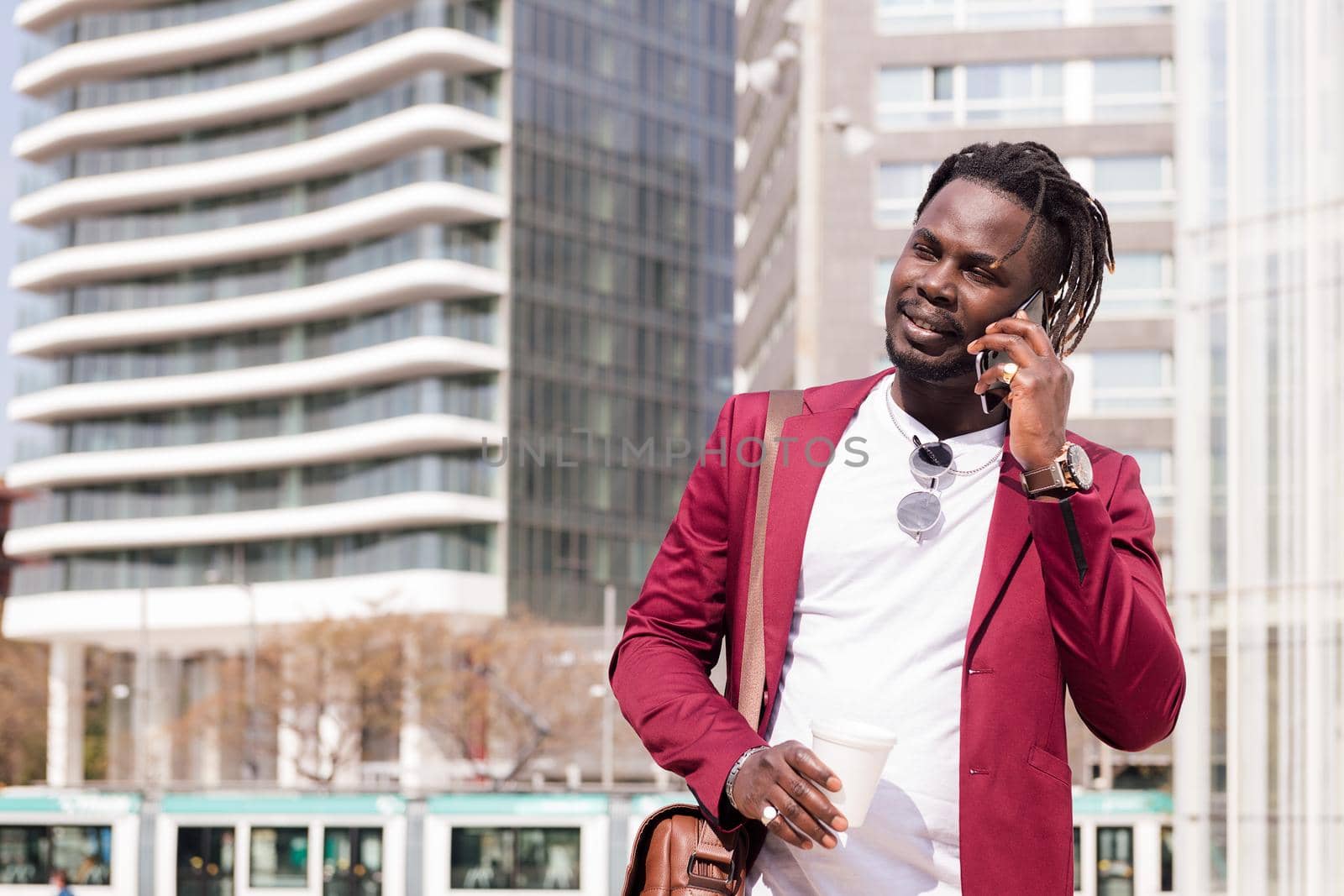 elegant black businessman walking through the city financial downtown talking on the phone with a coffee in his hand, concept of technology and communication, copy space for text