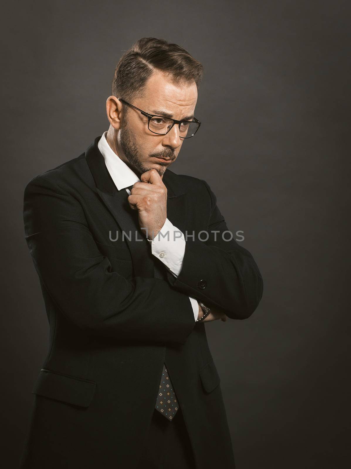 Doubting businessman crossed his arms touching his chin. Thoughtful intelligent man in glasses stands hesitantly looking to the side. Doubt concept. Toned image.