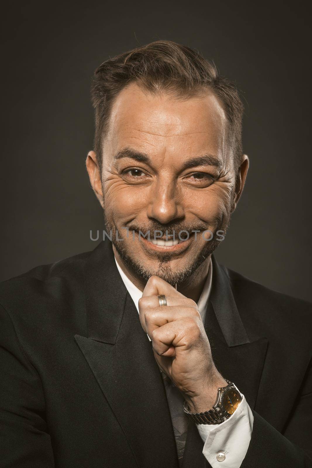 Attractive businessman charmingly smiling while looking at the camera and touching his beard with his hand. Close up portrait of mid aged caucasian man. Toned image by LipikStockMedia
