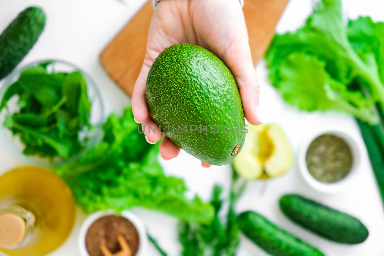 Woman cooking salad of fresh green vegetables and herbs. Cooking healthy diet or vegetarian food. Female hands hold avocado surrounded by green vegetables. Step by step recipe. Healthy eating. Ingredients for salad. Pattern flat lay with healthy vegetarian meal ingredients. Raw food concept. A variety of organic fruits and vegetables with avocado. Vegan menu. Fresh spinach and green onion leaves.