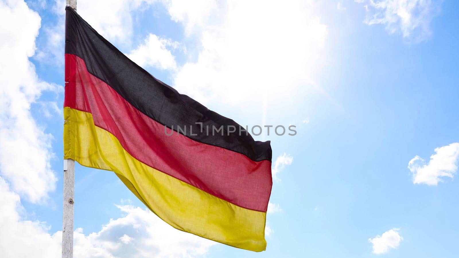 The official flag of Germany waving on a blue sky background. Horizontal banner design, with the German flag hanging on a sunny background with white clouds. Deutschland flag wide banner.