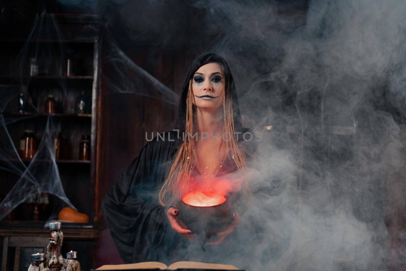 Halloween, witch use magic book and cauldron prepare poison or love potion. Black magic occult female wizard in dark room with smoke, spider web. Necromancer cooking magic potion. Close up