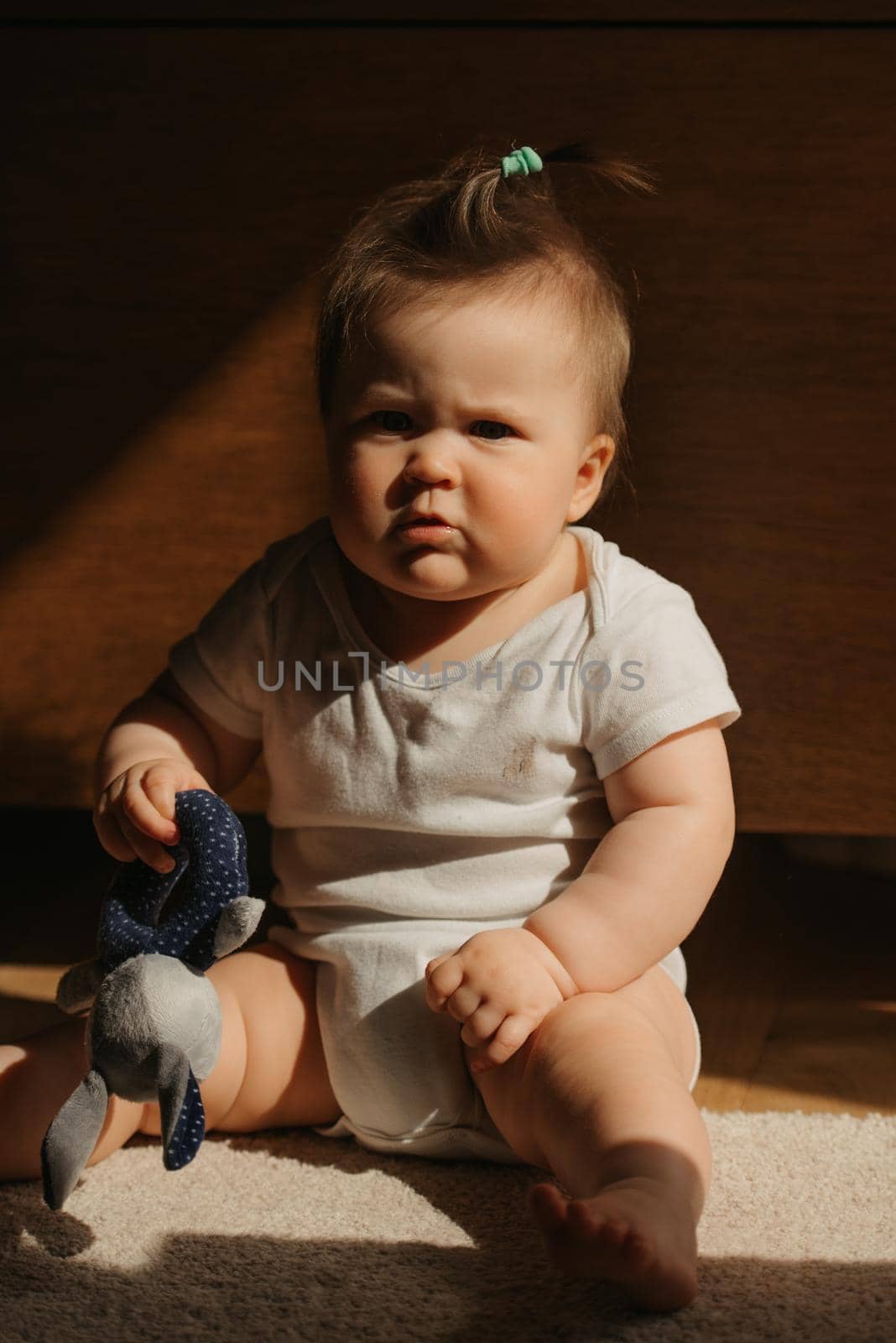A 7-month girl is sitting on the carpet dressed in a bodysuit at home. A cute infant is playing with a plush toy and squinting in the sunlight.