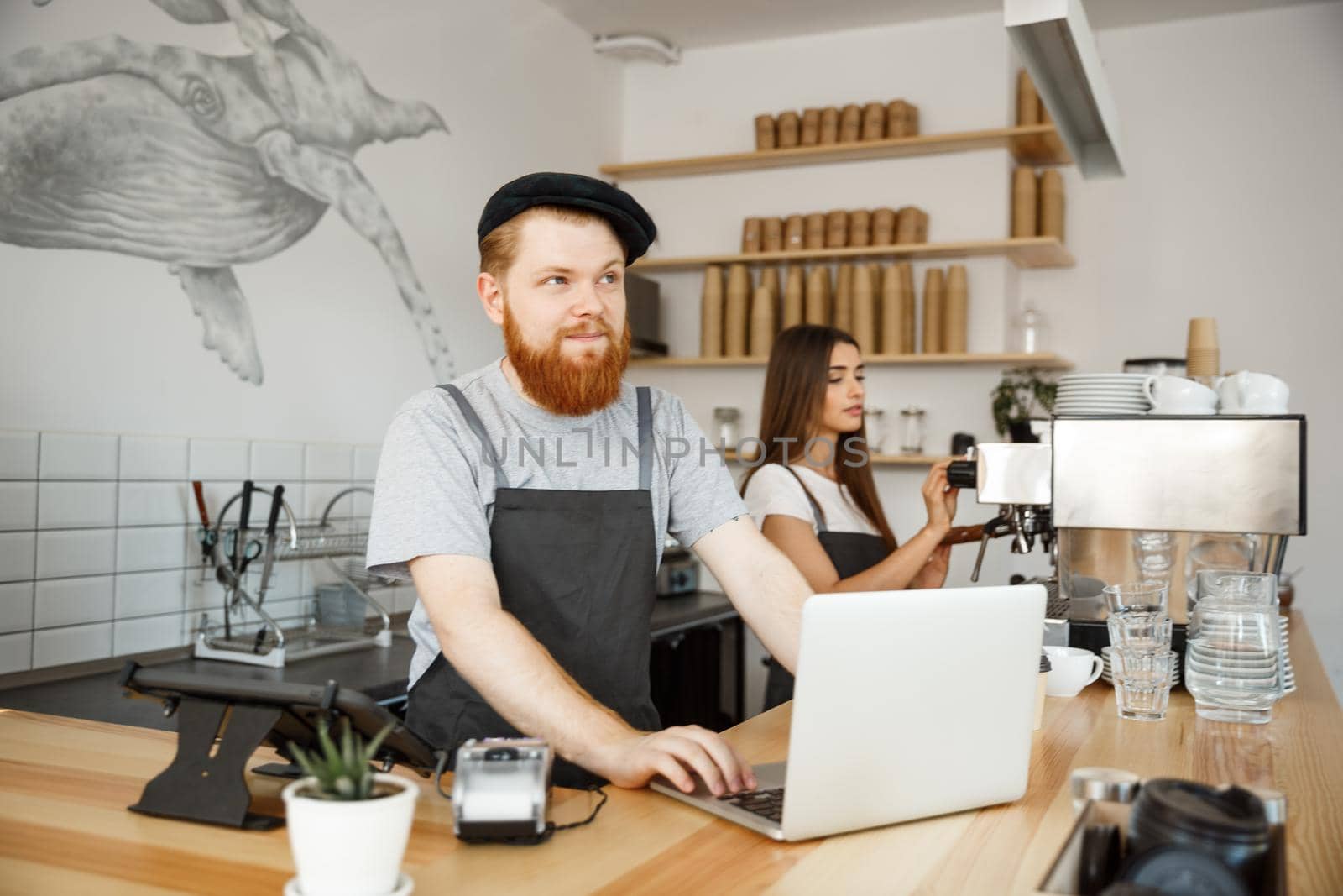 Coffee Business Concept - Young handsome bearded bartende, barista or manager working and planing in laptop at modern coffee shop.