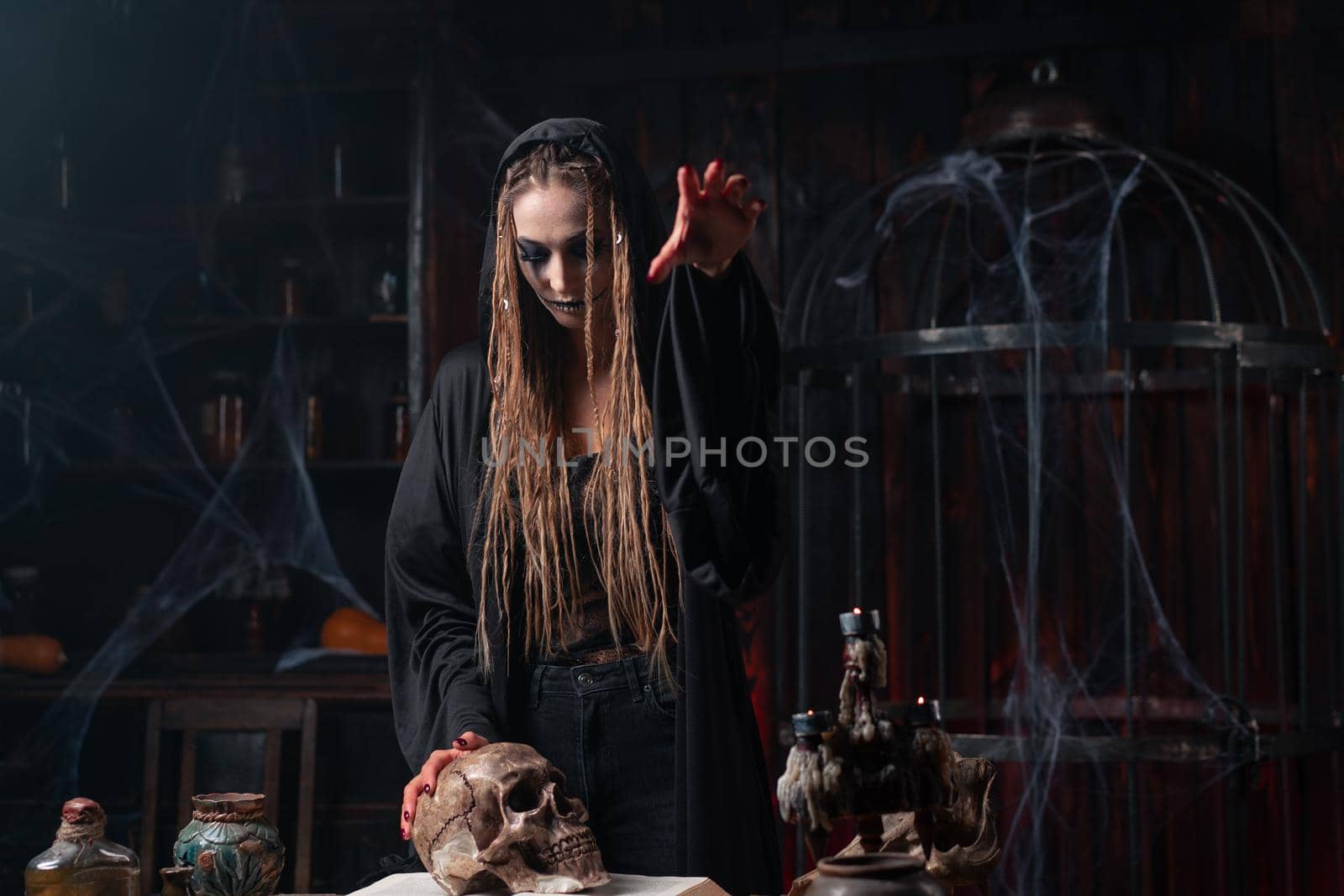 Halloween concept. Witch portrait close up with dreadlocks holding human skull in hand dressed black hood standing dark room side view making curse on camera Caucasian female necromancer