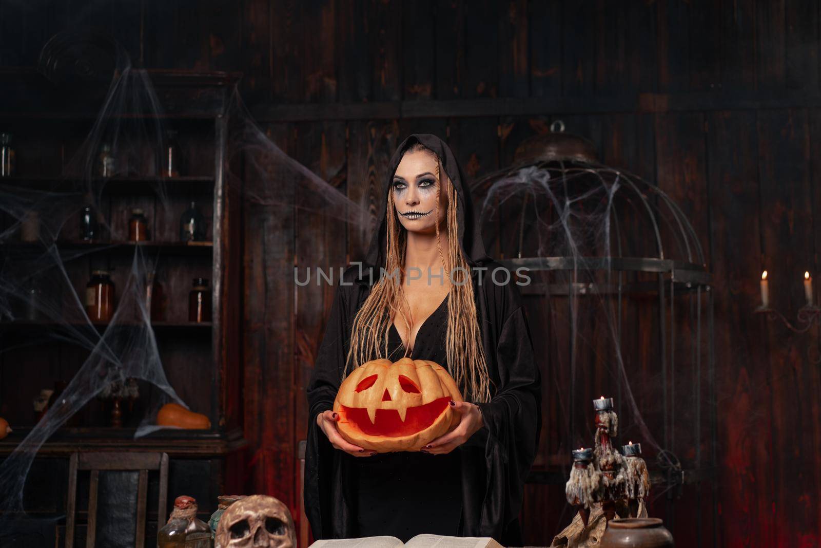 Halloween concept. Black witch holding Halloween pumpkin in hand with carved smily face in hand standing dark room. Female necromancer, dungeon place. Jack o lantern head. Wizard looking away