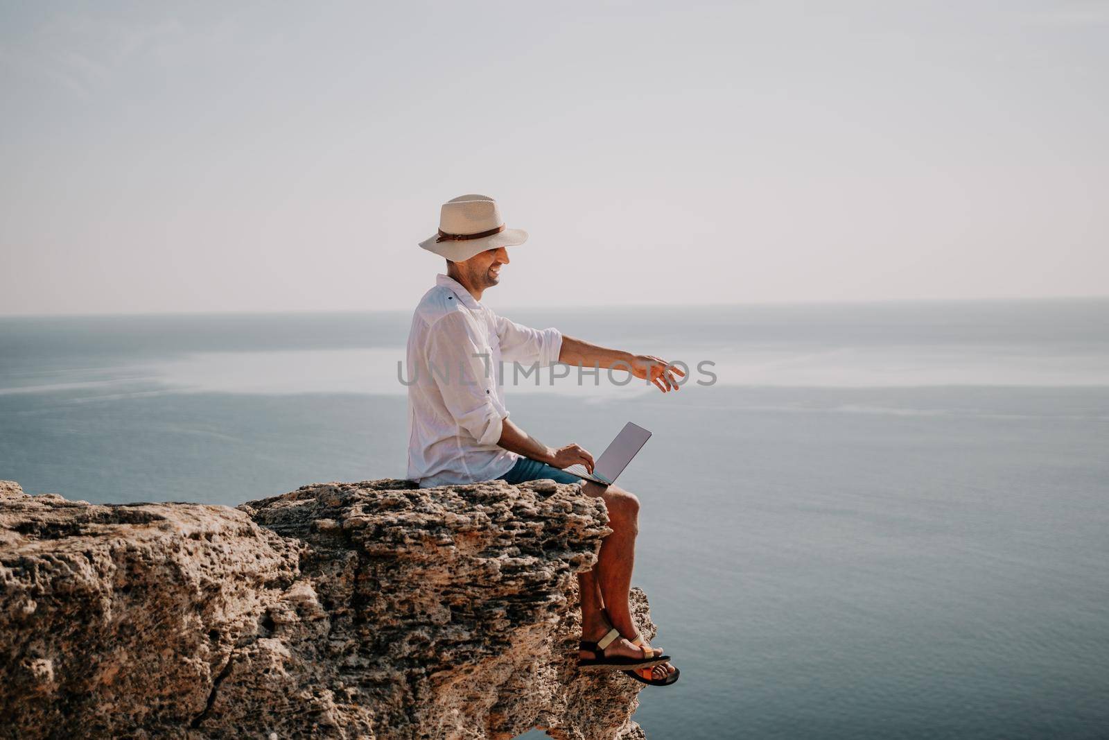 Digital nomad, man in the hat, a businessman with a laptop sits on the rocks by the sea during sunset, makes a business transaction online from a distance. Remote work on vacation. by panophotograph