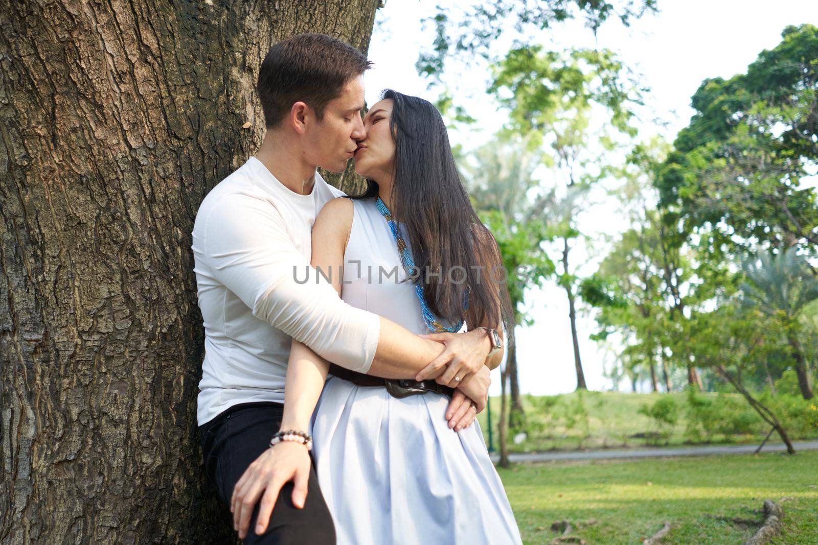 Pofile of a multiethnic newlywed couple kissing while leaning on a tree in a park