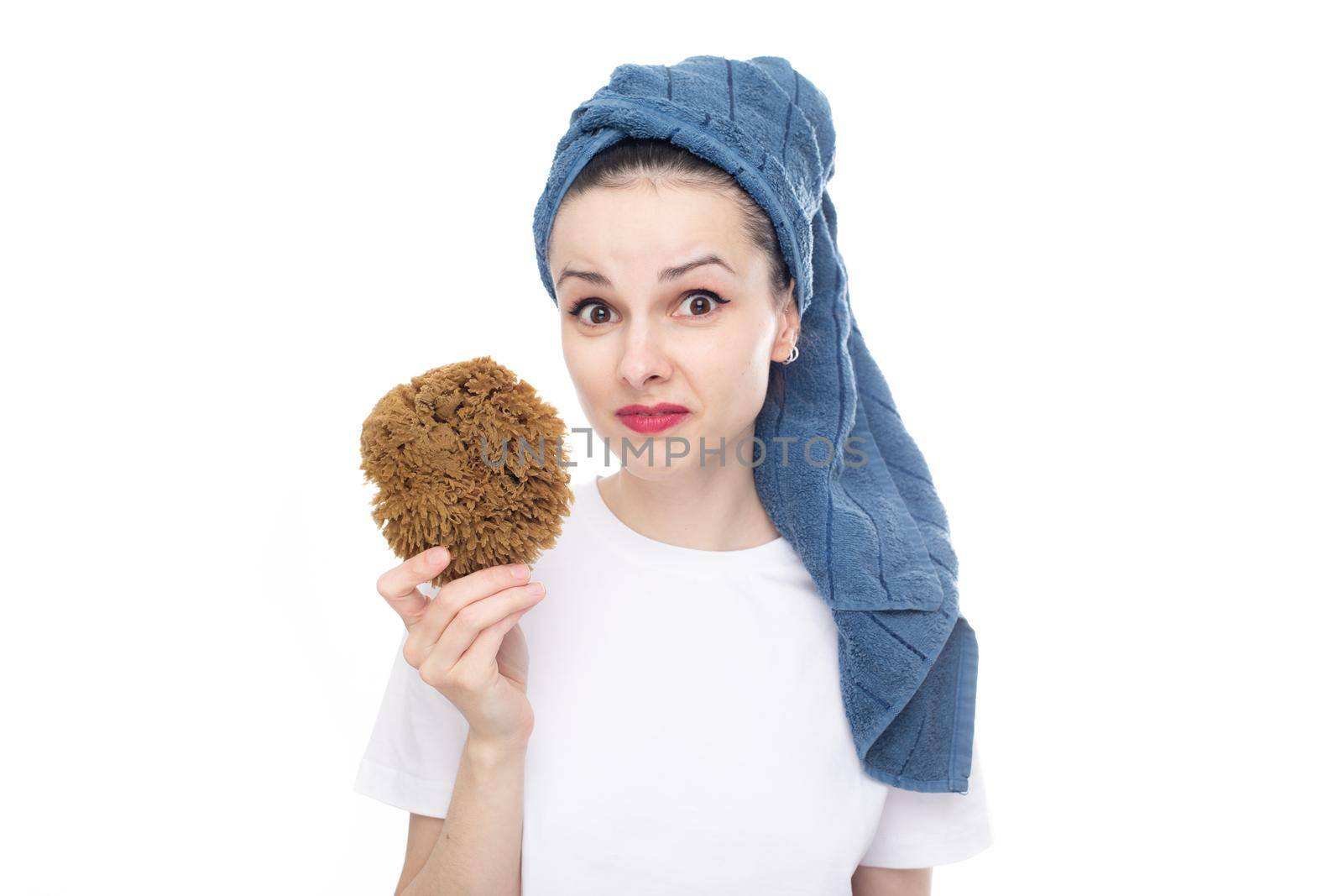 surprised woman in a white t-shirt and a blue towel on her head holds a body washcloth made of natural seaweed in her hand, white studio background. High quality photo