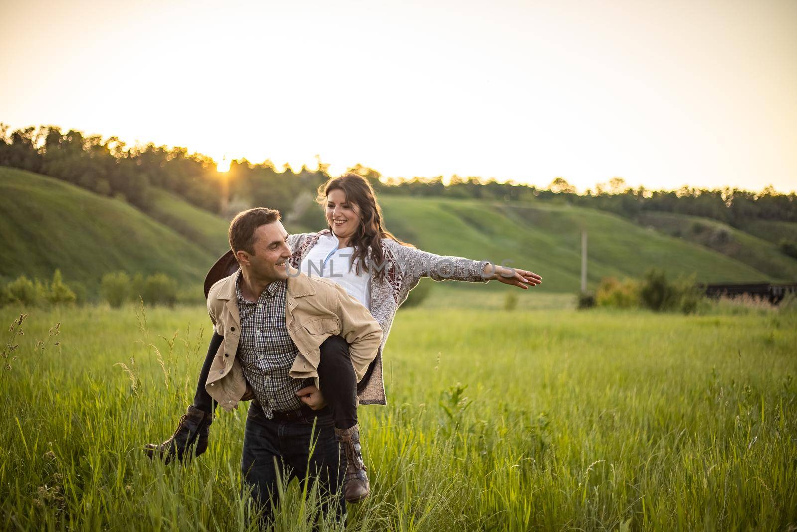young beautiful, happy couple on a green meadow in the rays of the setting sun, a man holds a girl on his back. cowboy clothes.rustic style by Anyatachka