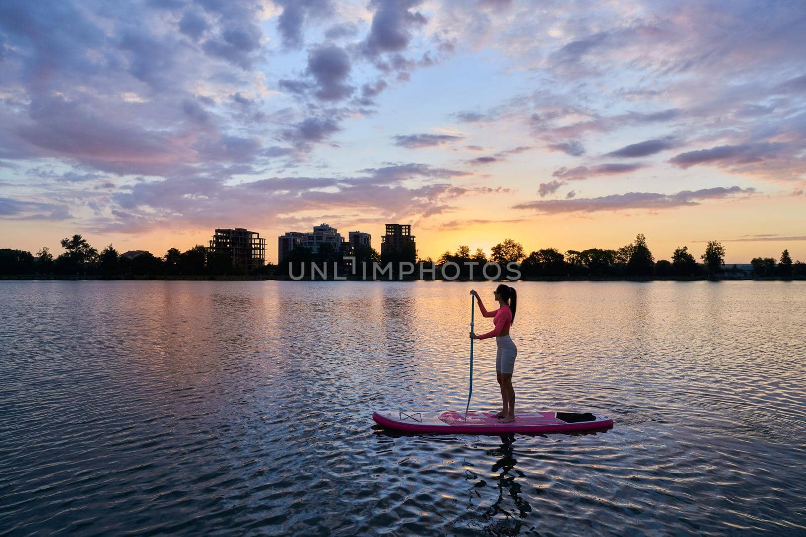Caucasian woman with slender body training on sup board during summer sunset. Young brunette enjoying evening time during water activity.