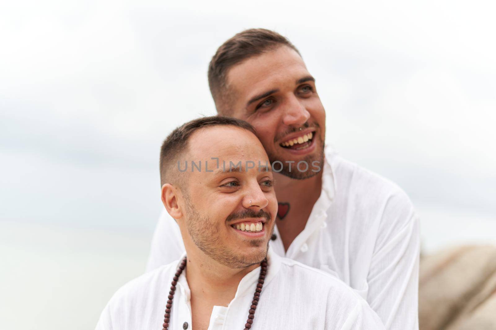 Close up portrait of a gay couple in white smiling as they look to the side embracing on the beach