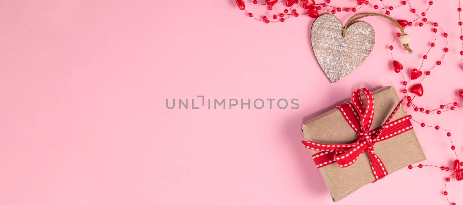 banner with One wooden heart and gift with red ribbon near beads with hearts on pastel pink colors. Valentine's Day background. Valentine's Day concept. Flat lay. Top view. by Leoschka