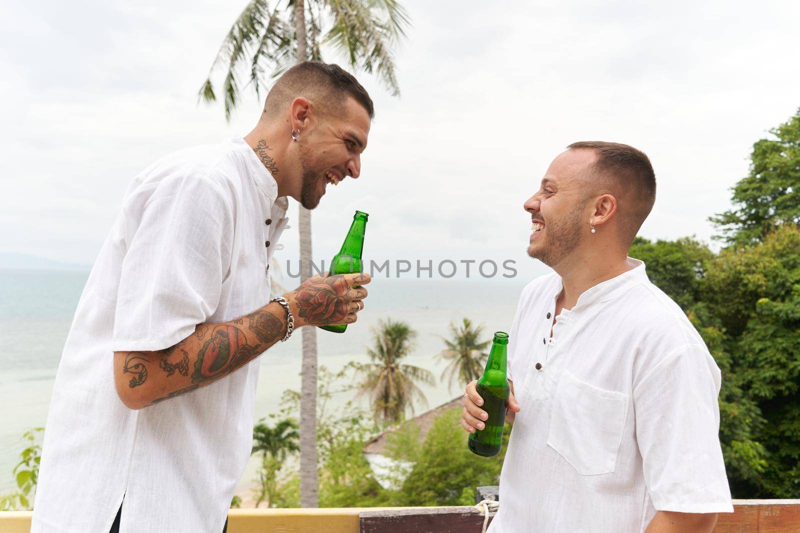 Homosexual couple drinking beer while laughing and talking next to a tropical beach