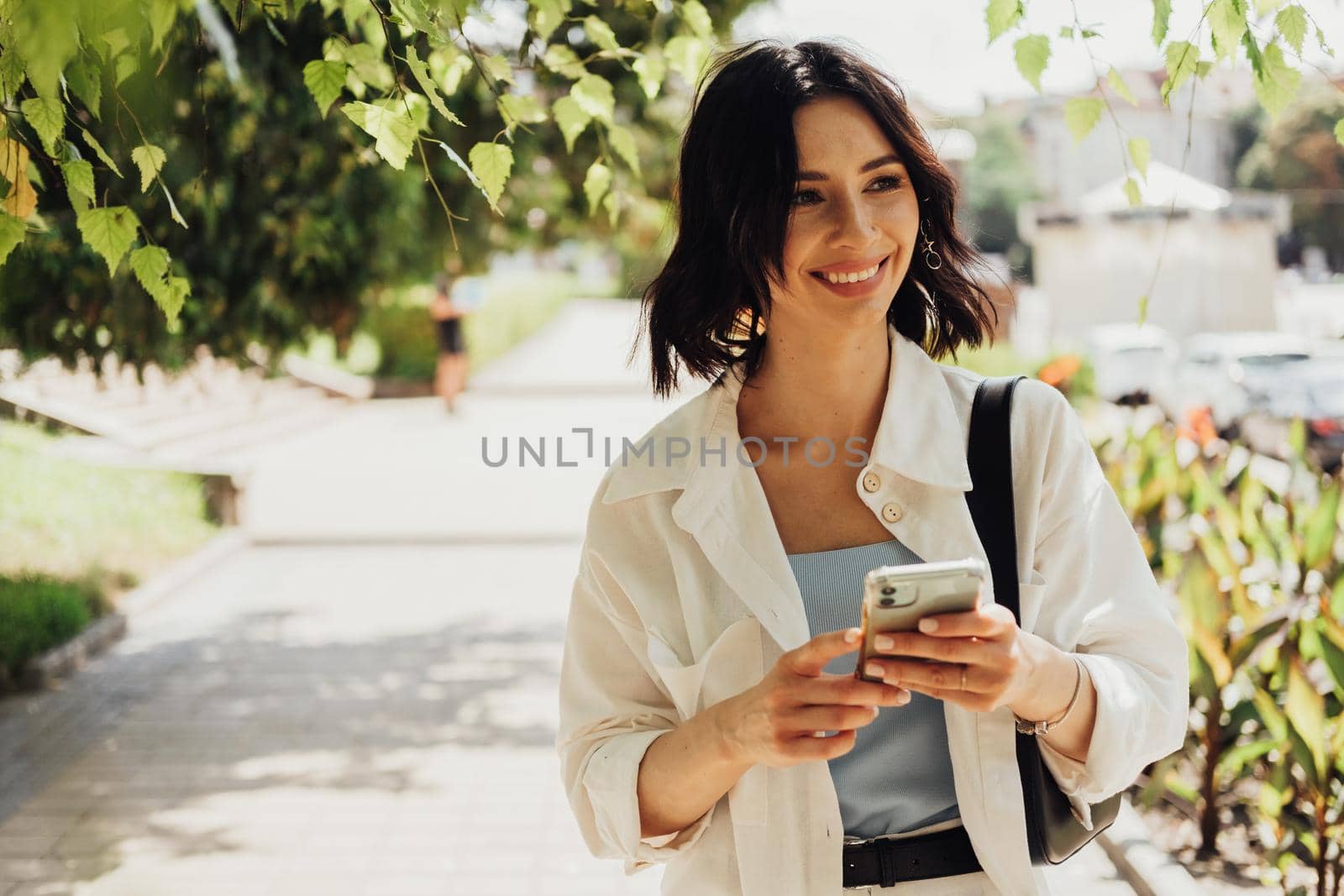Cheerful Young Woman Holding Smartphone While Walking Outdoors in Park at Summer Sunny Day, Copy Space