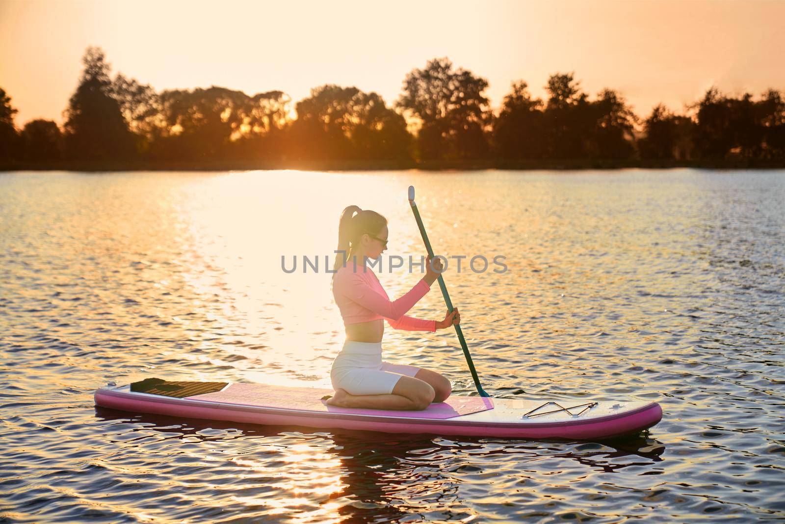 Attractive caucasian woman wearing colorful sport clothes and sunglasses rowing with paddle on sup board. Young lady with dark hair spending evening time for water activity.