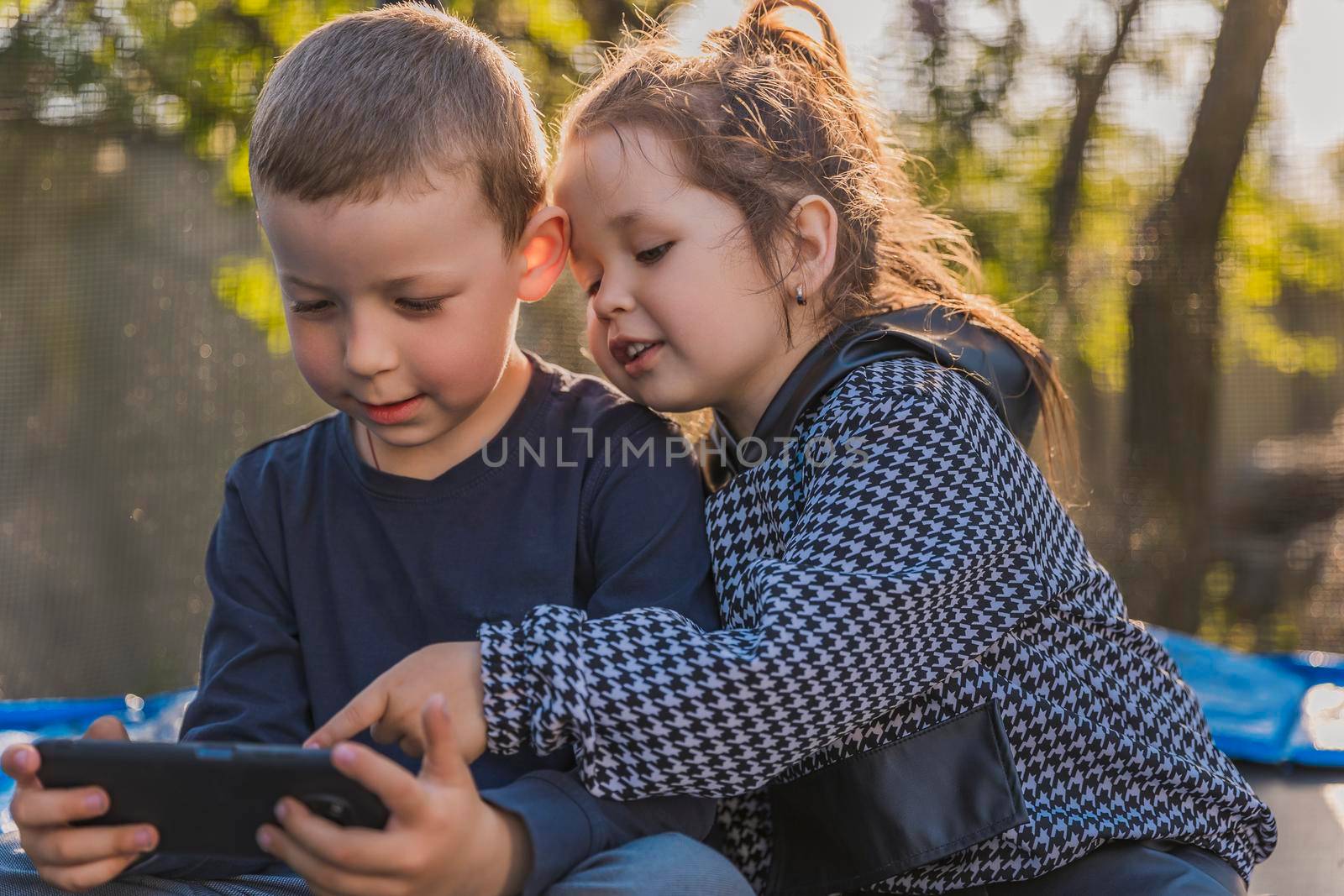 children look at the phone while sitting on a trampoline