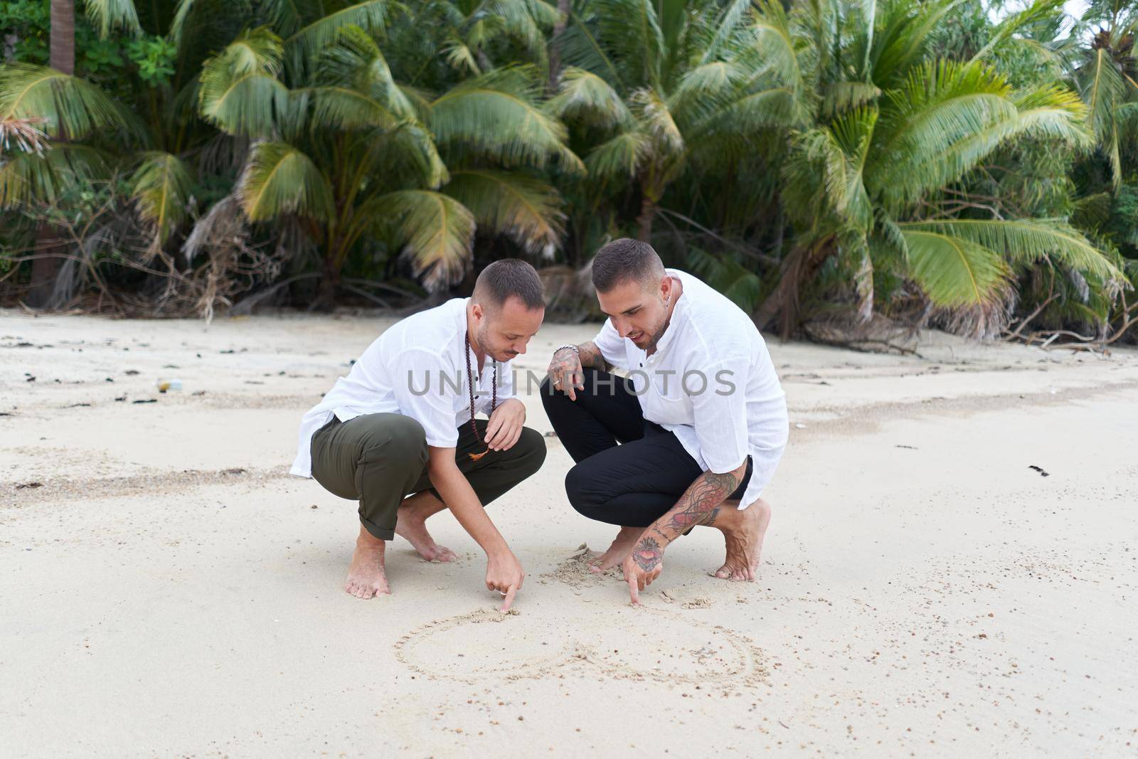 Homosexual couple crouching on a tropical beach drawing a heart in the sand together