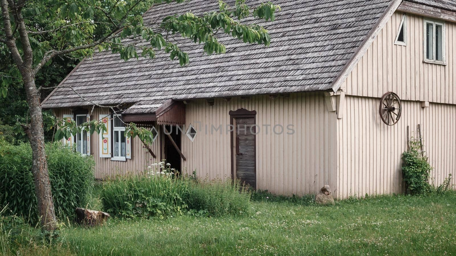 Old, abandoned beige farmhouse with trees, grass, blue sky and a wagon wheel on the wall, Plateliai Lithuania.